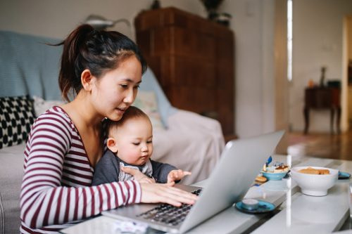 Photograph of a woman sitting at home working on a laptop computer with her baby on her lap, illustrating the human context of work.