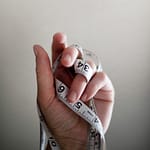 Photograph of a hand oriented vertically with the palm facing the camera and fingers folded with a tape measure entwined around the fingers.