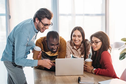 Photograph of four smiling co-workers collaborating around a laptop computer.
